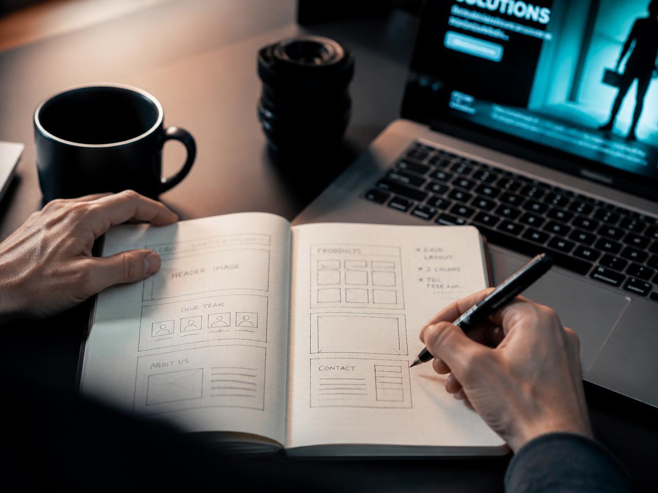 Hands sketching website wireframes in a notebook beside a laptop